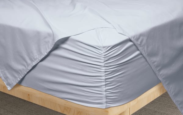 Light gray fitted sheet with ruched center detail on a wooden bed frame, paired with matching light gray flat sheets, showcasing smooth, soft fabric texture.