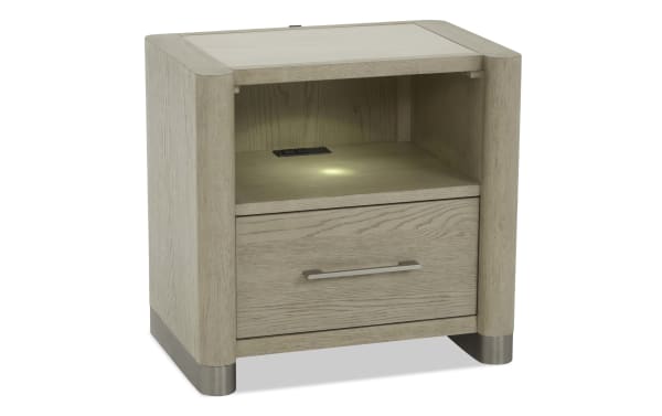 Light wood nightstand with a single drawer, open shelf, metal handle, metal base accents, and built-in power outlets for charging devices.