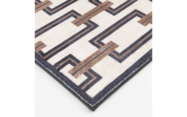 Corner of a modern area rug with a geometric pattern in beige, brown, and dark gray colors, featuring a soft textured surface and stitched edges.