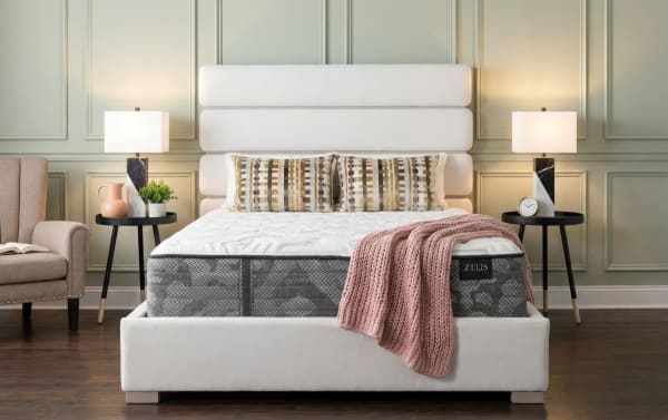 Modern bedroom setup featuring a Zeus brand mattress with a white upholstered bed frame and headboard, patterned pillows, a pink knitted throw, black marble table lamps, and wooden flooring.