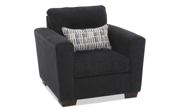 Dark gray fabric armchair with wide padded arms, a cushioned backrest, and a patterned lumbar pillow, featuring wooden block feet.