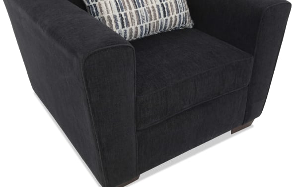 Dark charcoal gray upholstered armchair with wide padded arms, plush seat cushion, wooden block feet, and a patterned accent pillow.