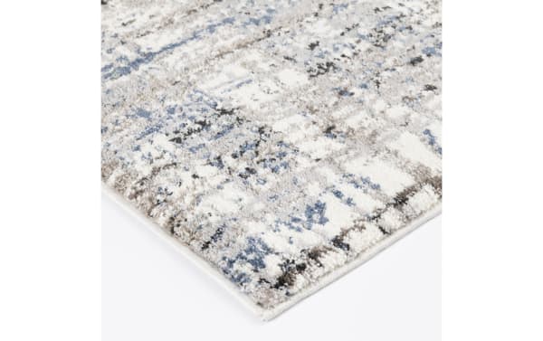 Corner close-up of a soft, plush area rug with a distressed pattern in shades of blue, gray, and white, featuring a thick pile and neatly finished edges.