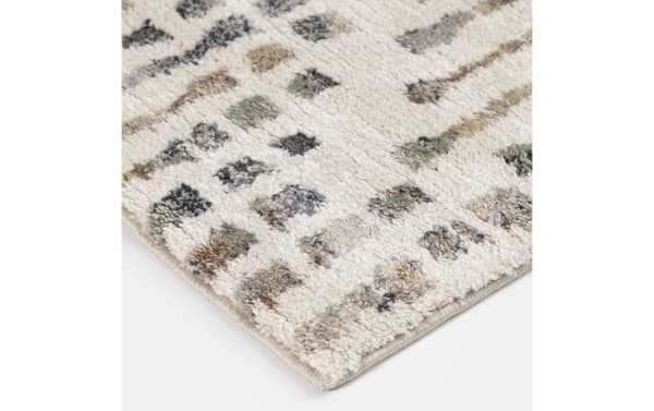 Close-up of a soft, plush area rug corner with a beige base and multicolored geometric pattern in gray, brown, and green tones.