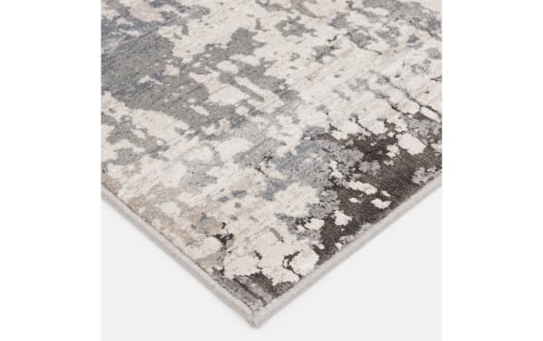 Corner view of a modern area rug with an abstract pattern in shades of gray, beige, and cream, featuring a soft, plush texture and neatly finished edges.
