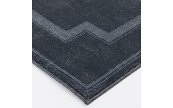 Dark gray corner of a soft, plush area rug with a subtle geometric border pattern in lighter gray tones, featuring tightly woven edges.