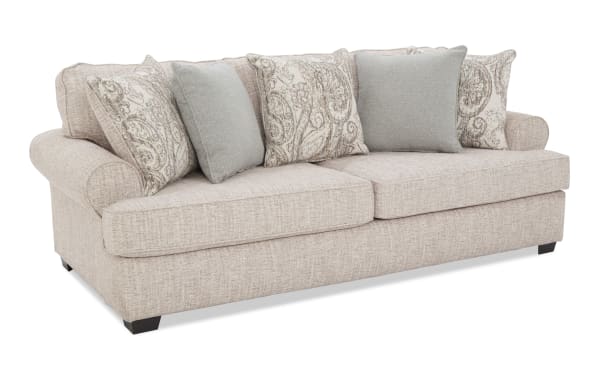 Beige fabric sofa with rolled arms and dark wooden legs, featuring five decorative cushions in light blue and paisley patterns, ideal for living room seating.