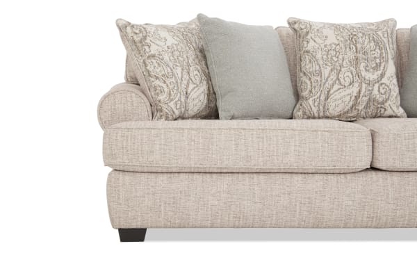 Beige fabric sofa with rolled armrest, featuring textured upholstery and decorative throw pillows in paisley and solid patterns, ideal for living room decor.