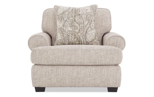 Beige fabric armchair with rounded arms and dark wooden legs, featuring a decorative beige and gray patterned throw pillow.