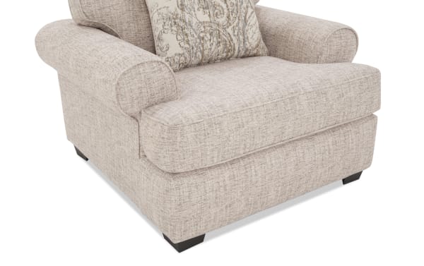 Beige fabric armchair with rolled arms, thick cushioned seat, dark wooden legs, and a decorative patterned throw pillow.