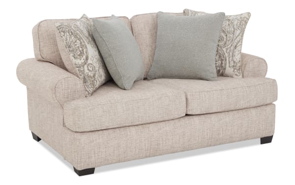 Beige fabric loveseat with rolled arms, dark wooden legs, and four decorative throw pillows in light blue and patterned beige.