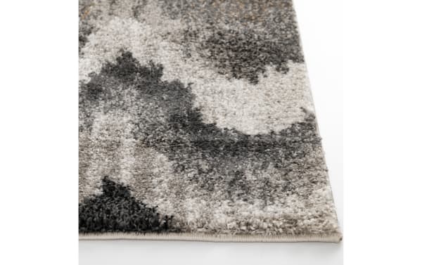 Close-up of a plush area rug corner with a soft, dense pile in a modern abstract pattern of gray, black, and beige tones, highlighting texture and edge binding.