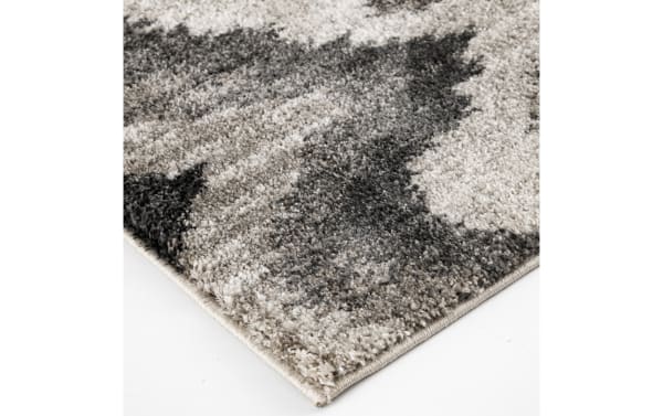 Corner close-up of a plush, high-pile area rug with a modern abstract pattern in shades of gray, black, and beige on a white floor.