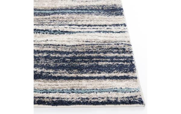 Area rug corner with a textured, striped pattern in shades of blue, beige, and light teal, featuring a soft, plush pile and tightly woven edges.