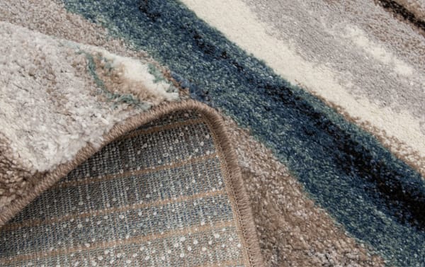 Close-up of a folded plush area rug with a textured, multicolored pattern featuring beige, cream, blue, and black stripes, highlighting its thick pile and woven backing.
