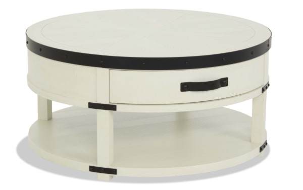 Round white wooden coffee table with black metal accents, a lower shelf for storage, and a drawer with a black handle, modern farmhouse style.