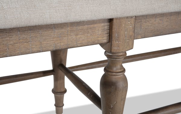 Light brown wood leg detail of Cottage Chic Light Brown & Beige Upholstered Dining Bench with weathered finish and beige polyester fabric upholstery.