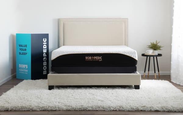 Copper Sport Gel 11.5'' Full Firm Low Profile Mattress Set by Bob-O-Pedic in black and white with copper accents, featuring temperature-regulating copper gel memory foam and a cool-to-touch cover, displayed on a beige upholstered bed frame with a plush white rug and modern black side table.