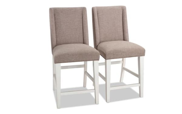 Set of 2 Montana Dining rustic counter height stools with beige polyester fabric upholstery, solid back, white rubberwood legs, and memory foam seats.