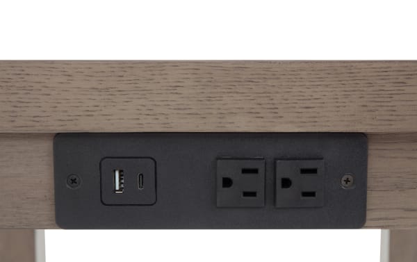 Black power outlet panel with two standard US electrical sockets and two USB ports (one USB-A and one USB-C) mounted on light brown wood surface.