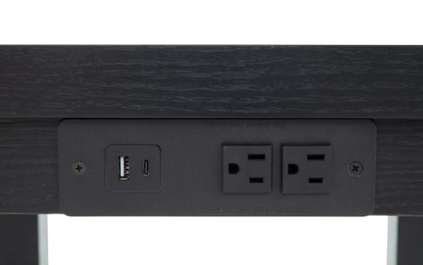 Black wooden desk with built-in power outlet panel featuring two standard US electrical sockets and dual USB ports (USB-A and USB-C) for charging devices.