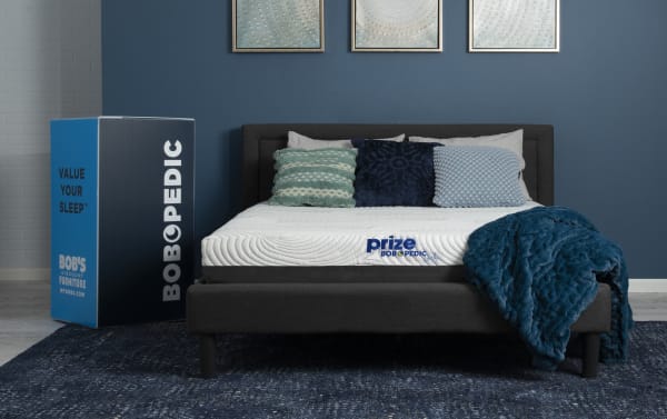 Prize Gel 10'' Full Firm Mattress from Bob-O-Pedic Prize collection with white quilted cover, gel memory foam, and boxed packaging shown on dark gray bed frame with blue pillows and blanket.