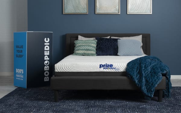 Power Bob Essential Base with Prize Gel 10'' Twin XL Firm Mattress from Bob-O-Pedic Prize collection, featuring white gel memory foam mattress with blue logo, dark gray adjustable base, blue textured throw, and decorative pillows on a blue rug.