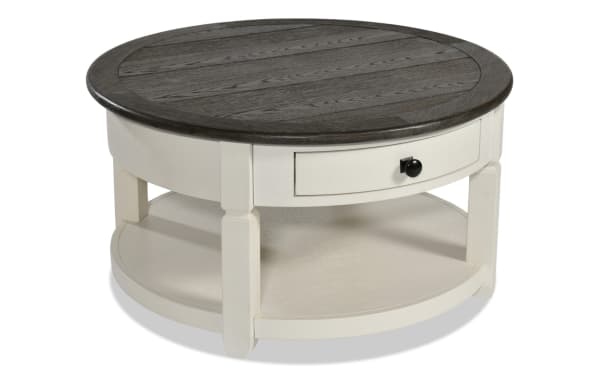 Round farmhouse-style Laurel Entertainment & Occasional coffee table with white MDF base, dark brown distressed wood top, and black hardware drawer for storage.