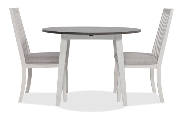 Blake 3 Piece White & Gray Upholstered Dining Set with two gray fabric polyester slat-back chairs and a round drop leaf table. Transitional style with MDF, hardwood, rubberwood, and birch veneers. Ideal for small spaces and apartments.