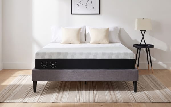 12'' Foam Queen Firm Mattress with white and gray tight-top design, firm foam support, shown on gray upholstered bed frame with beige pillows and black side table. Adjustable base compatible for enhanced sleep comfort.