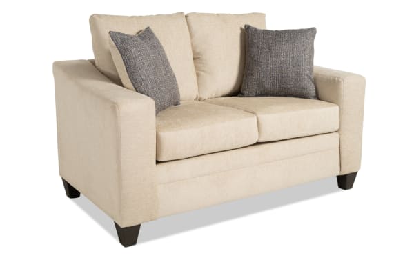 Claudia 62'' beige loveseat in transitional style with soft polyester fabric upholstery, featuring two gray textured accent pillows and dark tapered legs.