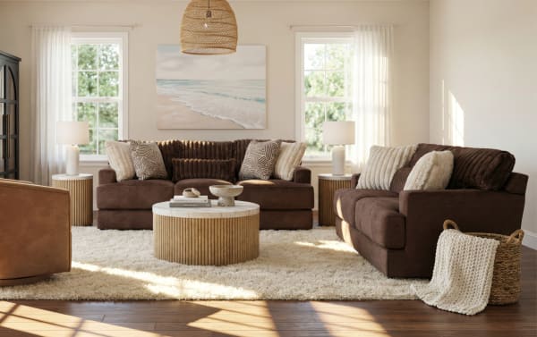 Living room with brown plush sofas, beige textured throw pillows, round wooden coffee table with marble top, cream shag rug, and natural light from windows.
