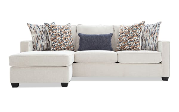 Modern beige fabric sectional sofa with left chaise, featuring textured upholstery, multiple patterned throw pillows, and a dark blue lumbar pillow.