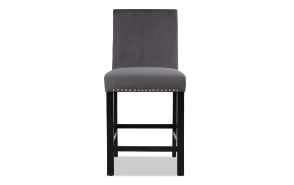 Gray upholstered bar stool with a high backrest, silver nailhead trim, and dark wooden legs with footrests, suitable for kitchen or bar seating.