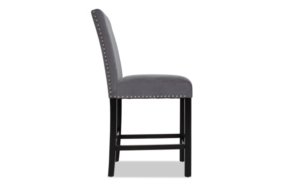 Gray velvet upholstered bar stool with silver nailhead trim, cushioned seat, and dark wooden legs with footrest, modern dining chair design.