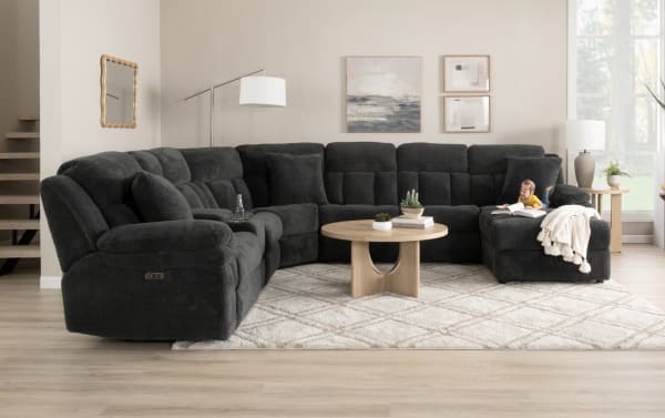 Dark gray sectional fabric sofa with plush cushioning and reclining seats, paired with a light wood round coffee table and neutral geometric rug in a modern living room.