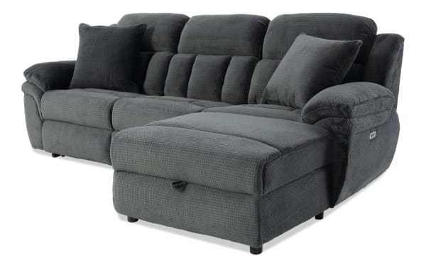 Dark gray plush fabric sectional sofa with chaise lounge, padded armrests, cushioned backrests, and two matching throw pillows.