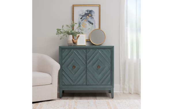 Blue-green wooden cabinet with diamond-patterned doors, brass knobs, and tapered legs, topped with a round gold-framed mirror, framed art, and a potted plant, set against a light wall with sheer curtains.