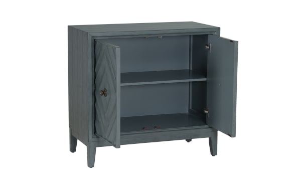 Blue-gray wooden cabinet with two open doors, featuring a chevron pattern on the front, one internal shelf, and brass knobs.