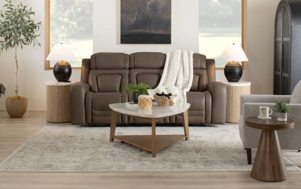 Modern living room with a brown leather sofa, white knitted throw, marble-top wooden coffee table, beige armchair, wooden side tables with black lamps, and a large area rug.