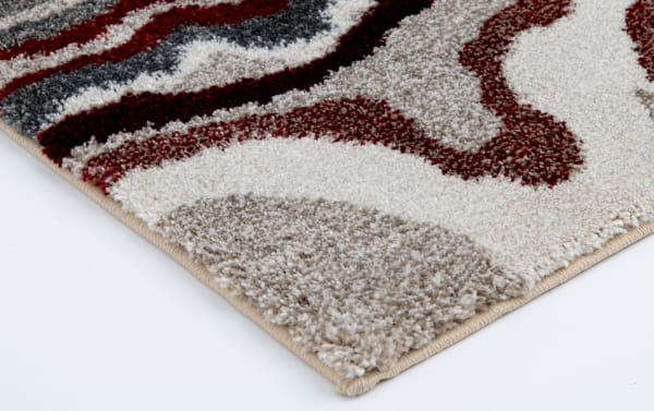 Close-up of a soft, plush area rug with a modern abstract pattern in beige, brown, cream, and red tones, showing detailed texture and edge stitching.