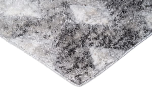 Close-up of a plush, shaggy area rug corner with a gray and white abstract pattern, highlighting soft texture and stitched edge.