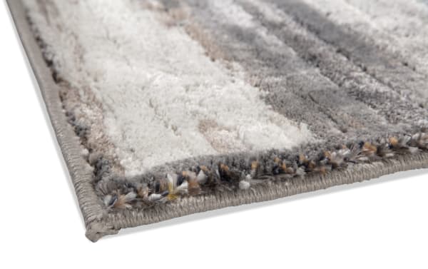 Close-up of a soft, plush area rug corner with a gray, white, and beige striped pattern and tightly stitched edges, highlighting texture and quality.