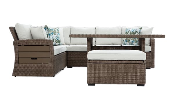 Outdoor sectional patio furniture set with brown wicker frame, beige cushions, and tropical leaf-patterned throw pillows, including a matching ottoman and rectangular table.