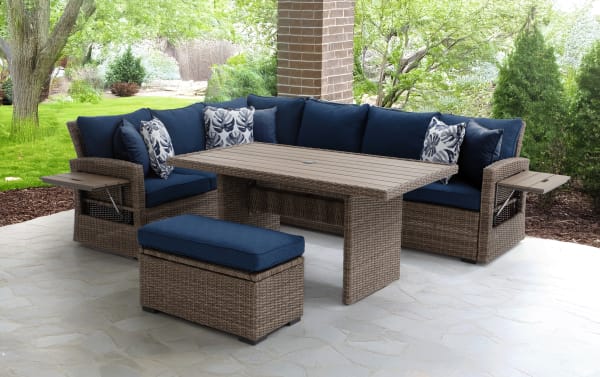 Outdoor patio furniture set with brown wicker frame, navy blue cushions, two floral throw pillows, a rectangular wicker table with slatted top, and a matching cushioned ottoman, ideal for garden or porch seating.