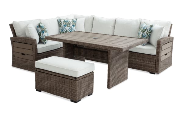 Outdoor sectional patio furniture set with brown wicker frame, beige cushions, tropical leaf-patterned throw pillows, and a matching ottoman with storage.