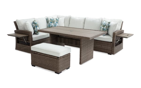 Outdoor patio furniture set with brown wicker frame, beige cushions, floral accent pillows, rectangular wood-look table, and matching cushioned ottoman.