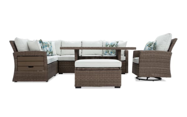 Outdoor patio furniture set with brown wicker frame, beige cushions, tropical leaf patterned throw pillows, including sectional sofa, swivel armchair, and ottoman with matching wicker table.