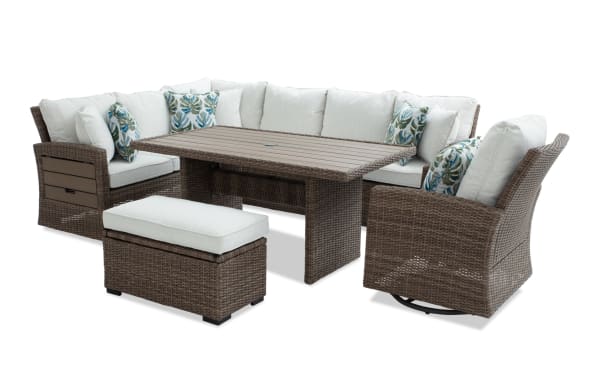 Outdoor patio furniture set featuring a brown wicker sectional sofa with beige cushions, a matching rectangular table, swivel chair, and cushioned ottoman with tropical leaf accent pillows.
