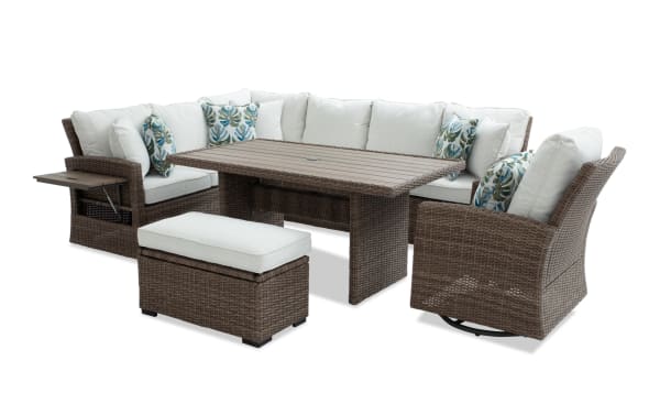 Outdoor patio furniture set with brown wicker frame, beige cushions, sectional sofa, swivel chair, ottoman, and rectangular dining table with side tray.